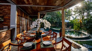 Uma área de refeições com uma mesa de madeira posta para quatro pessoas, rodeada por cadeiras, com vista para uma piscina e plantas tropicais.