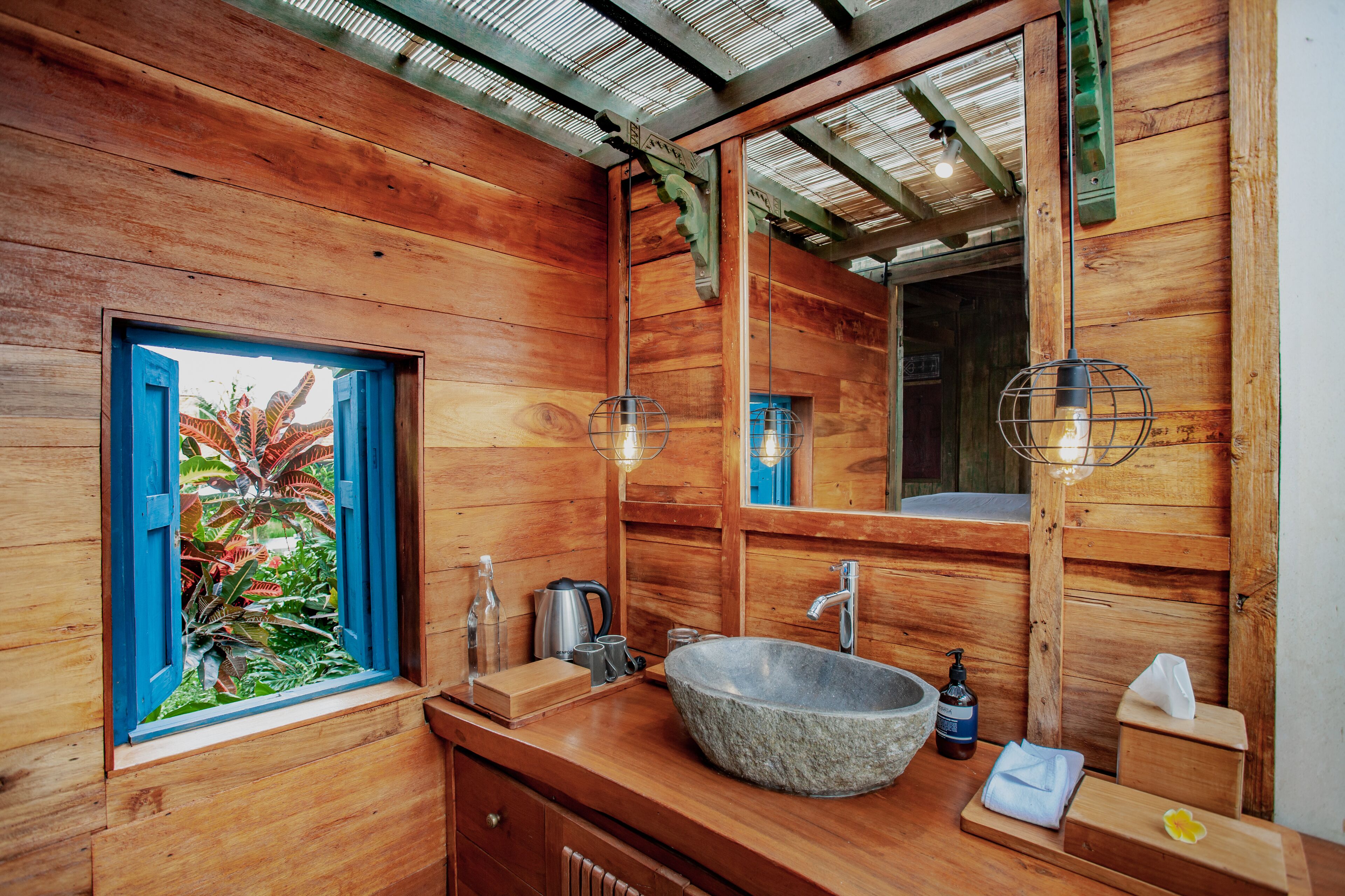 Une salle de bain en bois avec un grand lavabo en pierre, une fenêtre avec des volets bleus et un miroir.