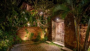 Vista noturna de um pátio com muro de tijolos, porta de madeira e vegetação exuberante.
