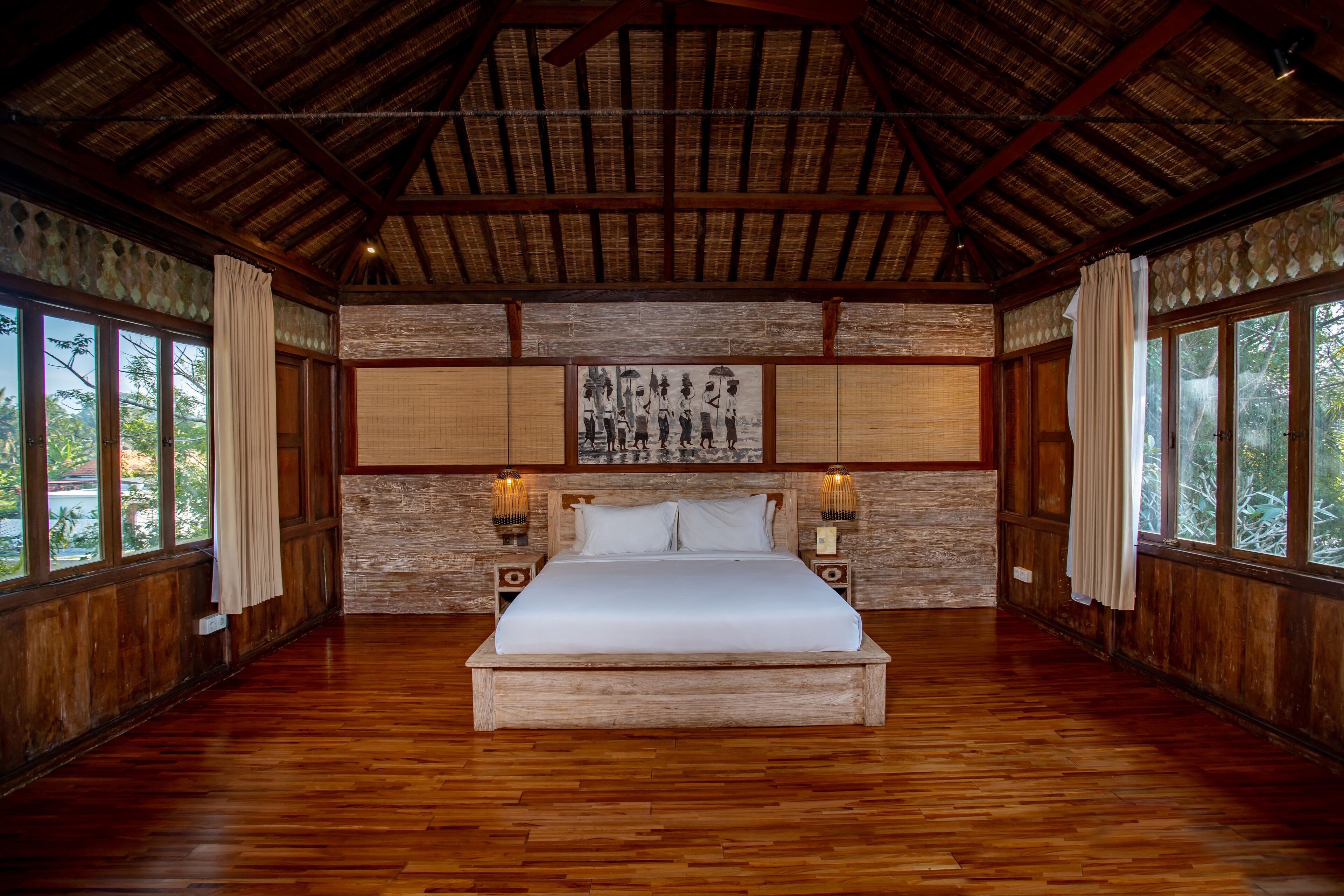 A large wooden-framed bedroom with a thatched roof, a bed centered in the room, and large windows on either side.