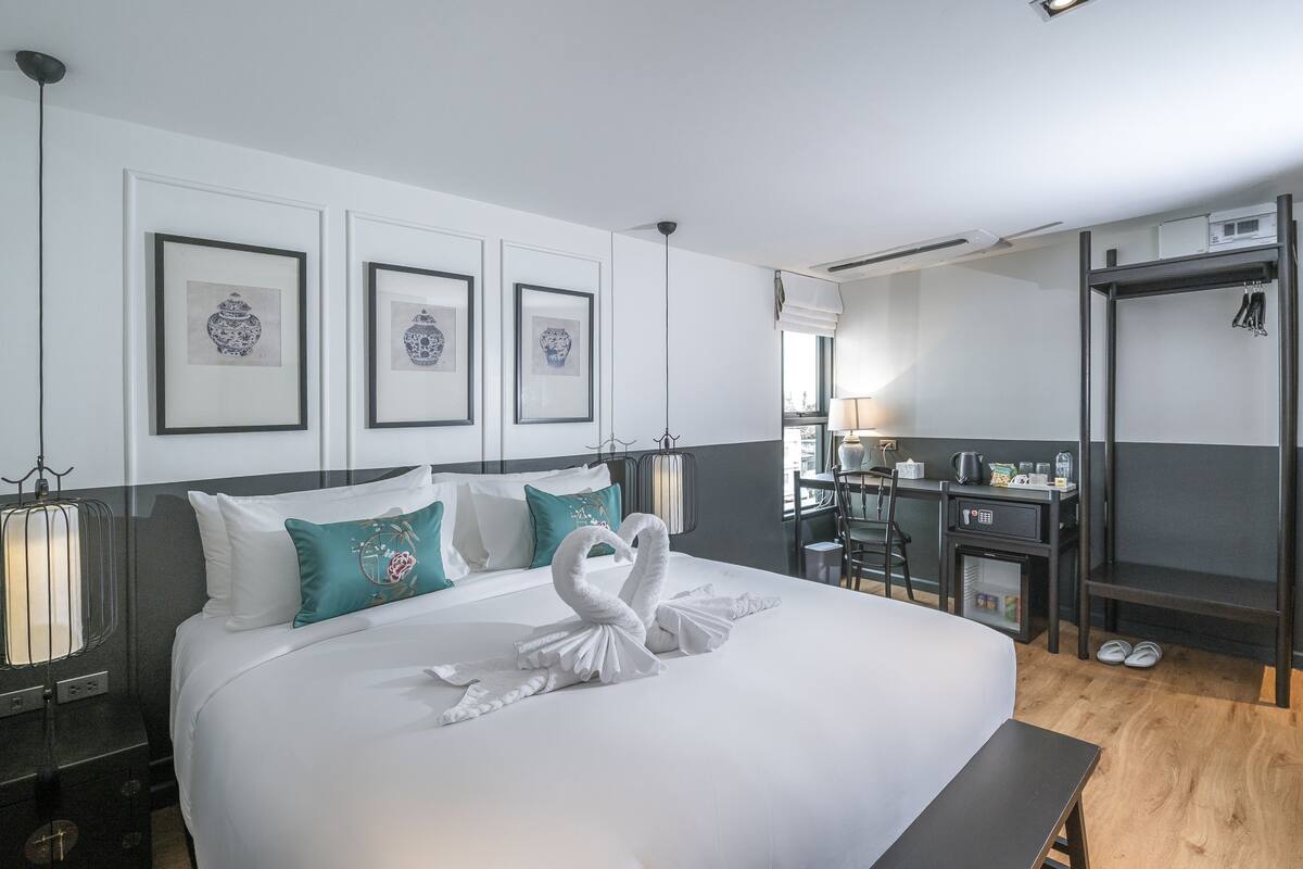 Une chambre d’hôtel avec un lit bien fait, un bureau et une chaise. Les murs sont ornés de tableaux encadrés et il y a une fenêtre avec vue.
