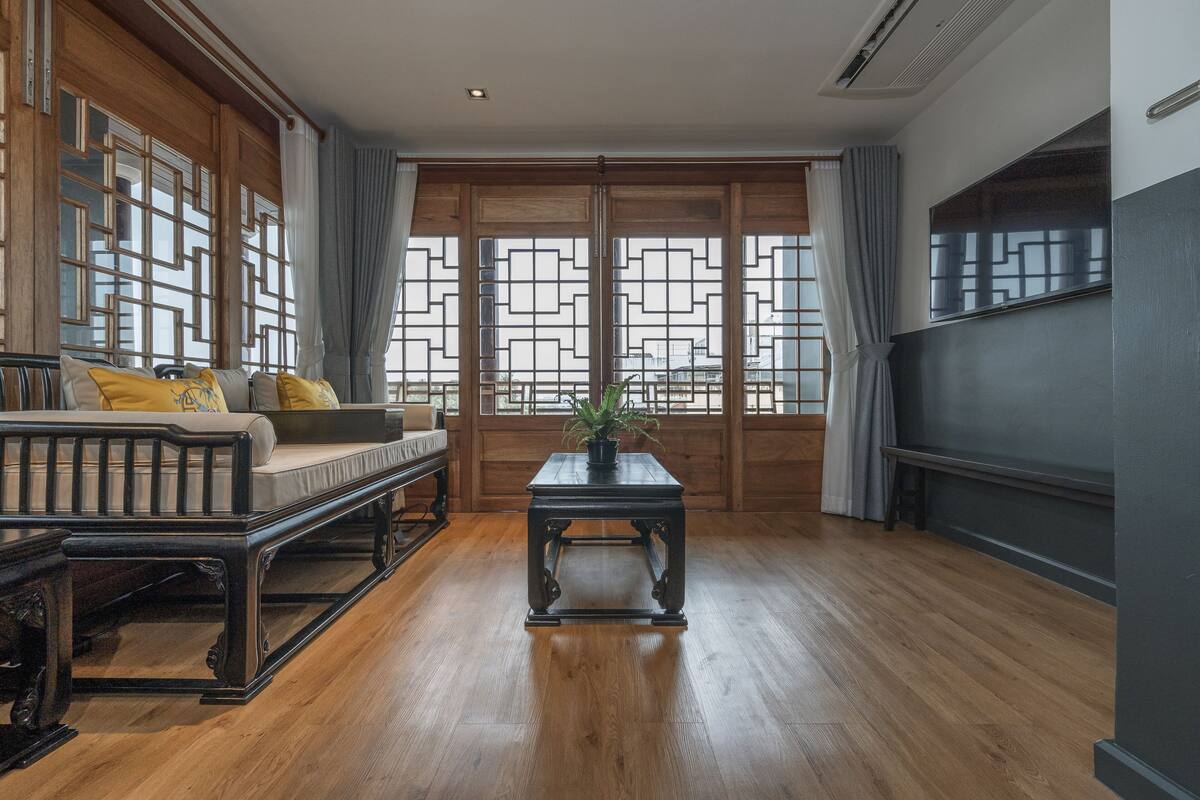 Une pièce à l’ambiance traditionnelle, avec un plancher en bois, un canapé agrémenté de coussins, une table ornée d’une plante et un téléviseur fixé au mur.