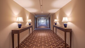 Pasillo de hotel con alfombra estampada, dos lámparas a cada lado y un largo corredor que lleva a una habitación luminosa al final.