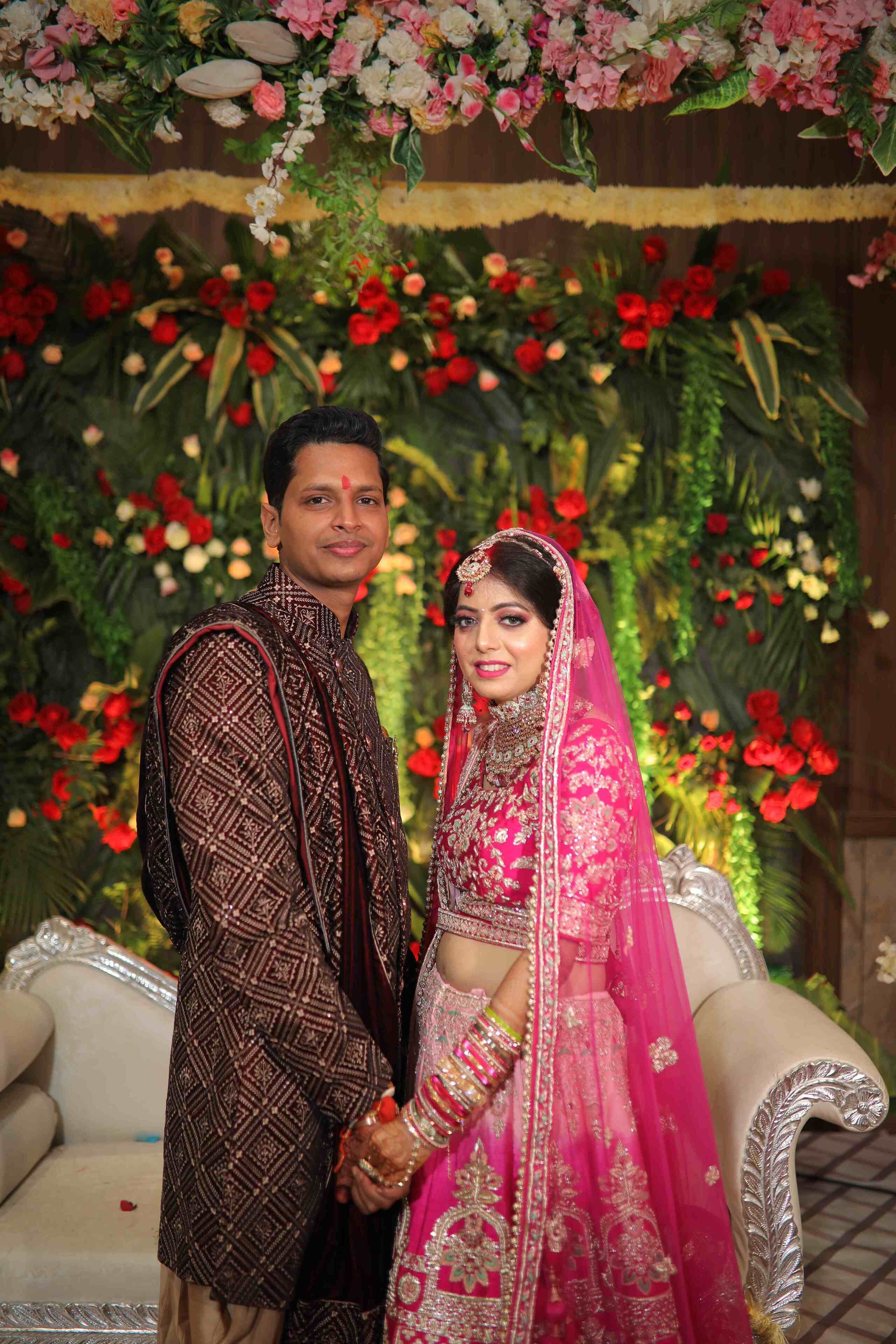 Una pareja vestida con trajes de novia tradicionales frente a un fondo floral.