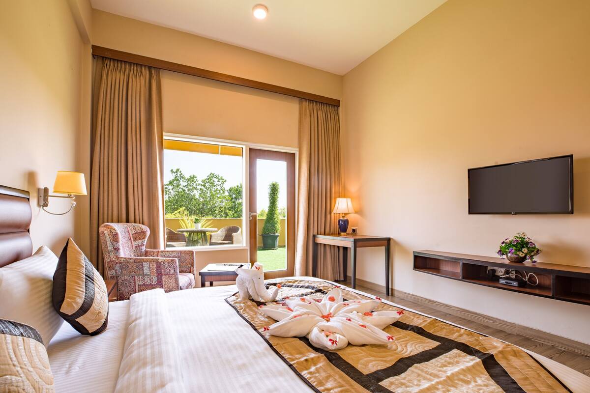 A hotel room with a large bed, a chair, a table, a TV, and a balcony with a view of greenery.