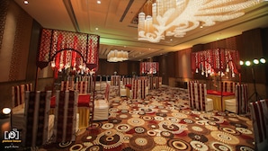 Un salón de banquetes con alfombra estampada, filas de mesas con manteles rojo y blanco, y candelabros ornamentados.
