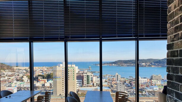 A dining area with tables and chairs, large windows with blinds, and a view of a coastal cityscape.