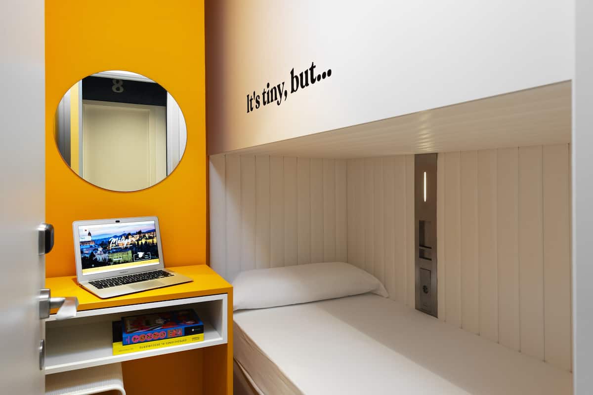 A compact room with a bed, a laptop on a small table, and a mirror on the wall.