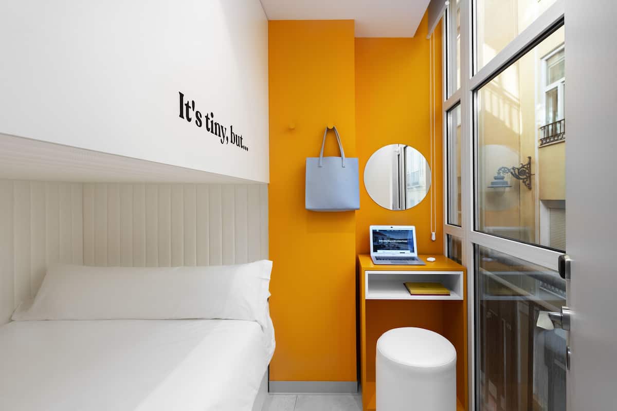 A modern hotel room with a bed, a desk with a laptop, a small round mirror, and a blue tote bag.