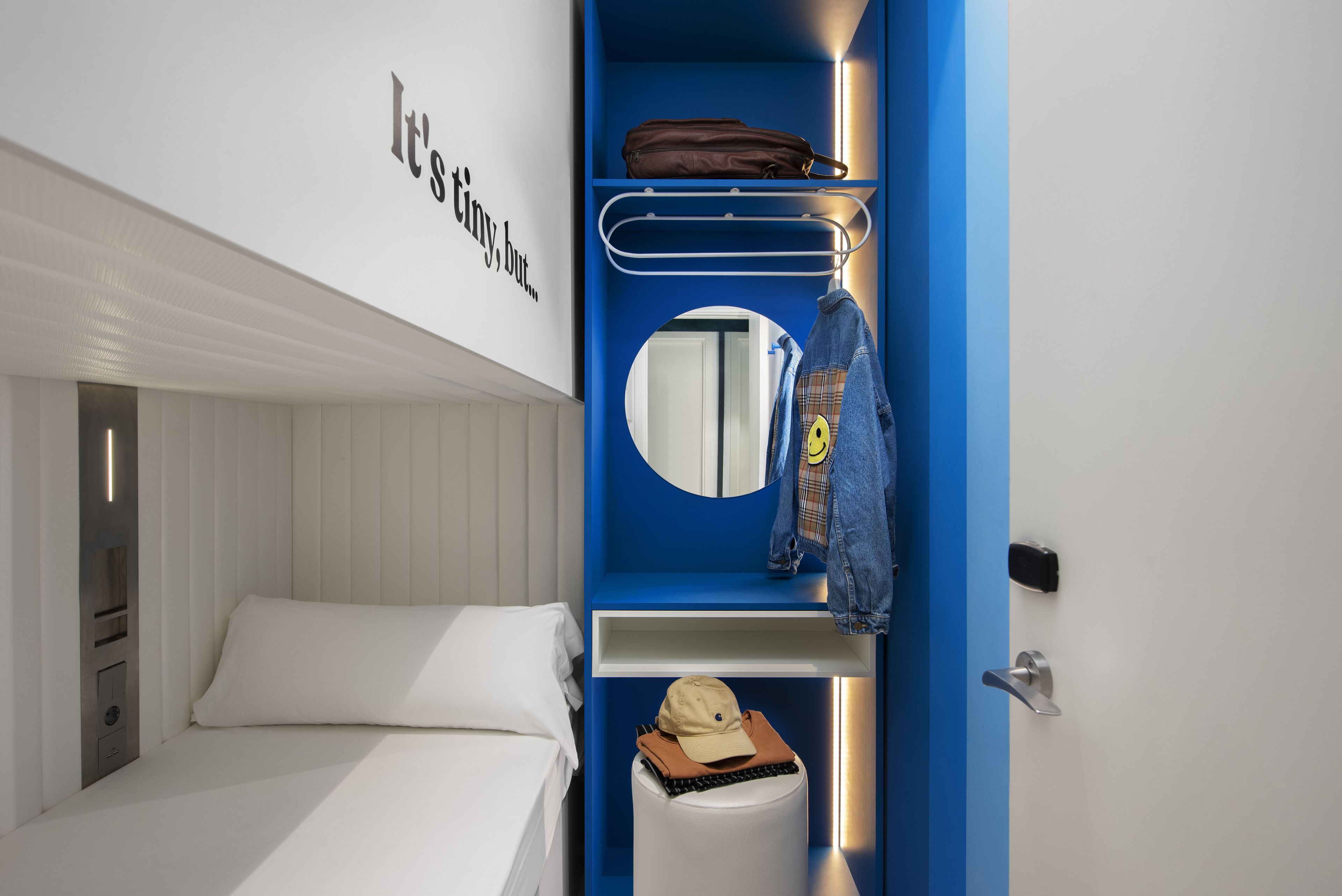A compact hotel room with a bunk bed, a small bedside table, a mirror, and a blue accent wall.