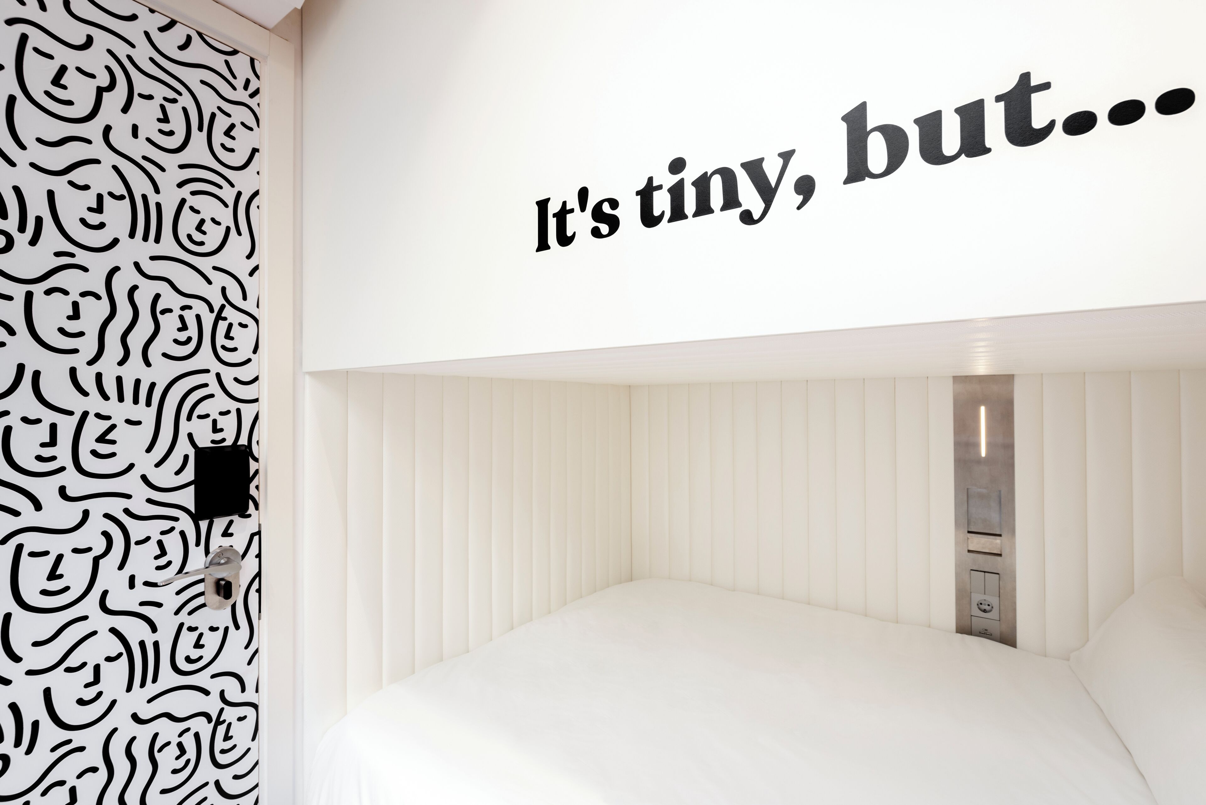 A room with a white bed, a wall with a black abstract pattern, and a text saying 'It's tiny, but...'.