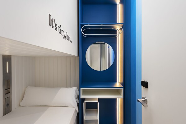 A bedroom with a bed, a wall-mounted shelf, a circular mirror, and a door with a handle.