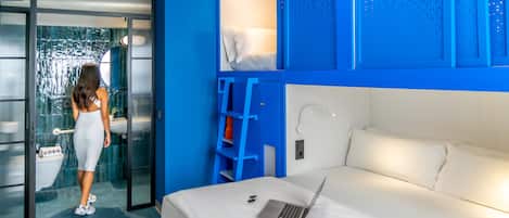 Quarto de hotel moderno com uma parede azul de destaque, uma cama branca, um laptop na cama e um banheiro com porta de vidro.