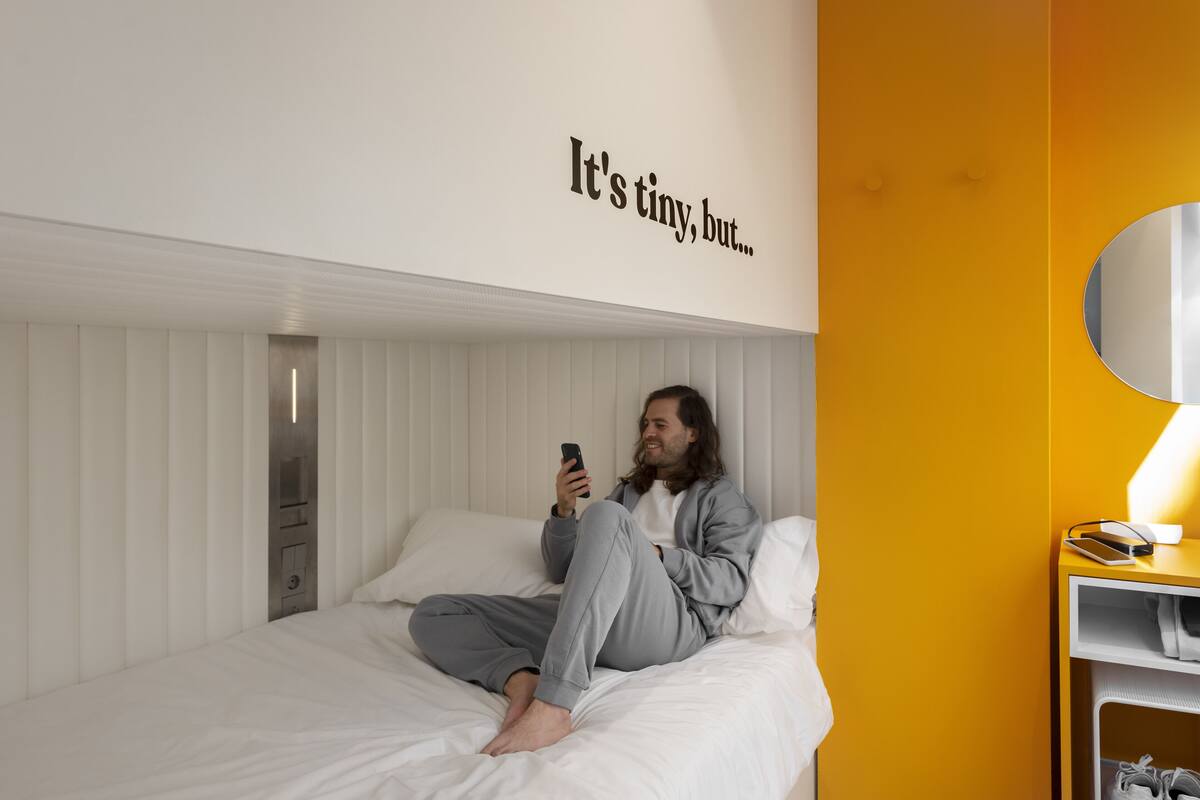 A person sitting on a bed in a room with a white and yellow color scheme, a wall with text, and a bedside table with a lamp and a phone.