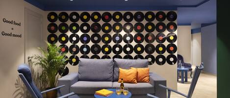 A modern living room with a wall of vinyl records, a grey sofa, blue chairs, and a small table with a bottle and glasses.