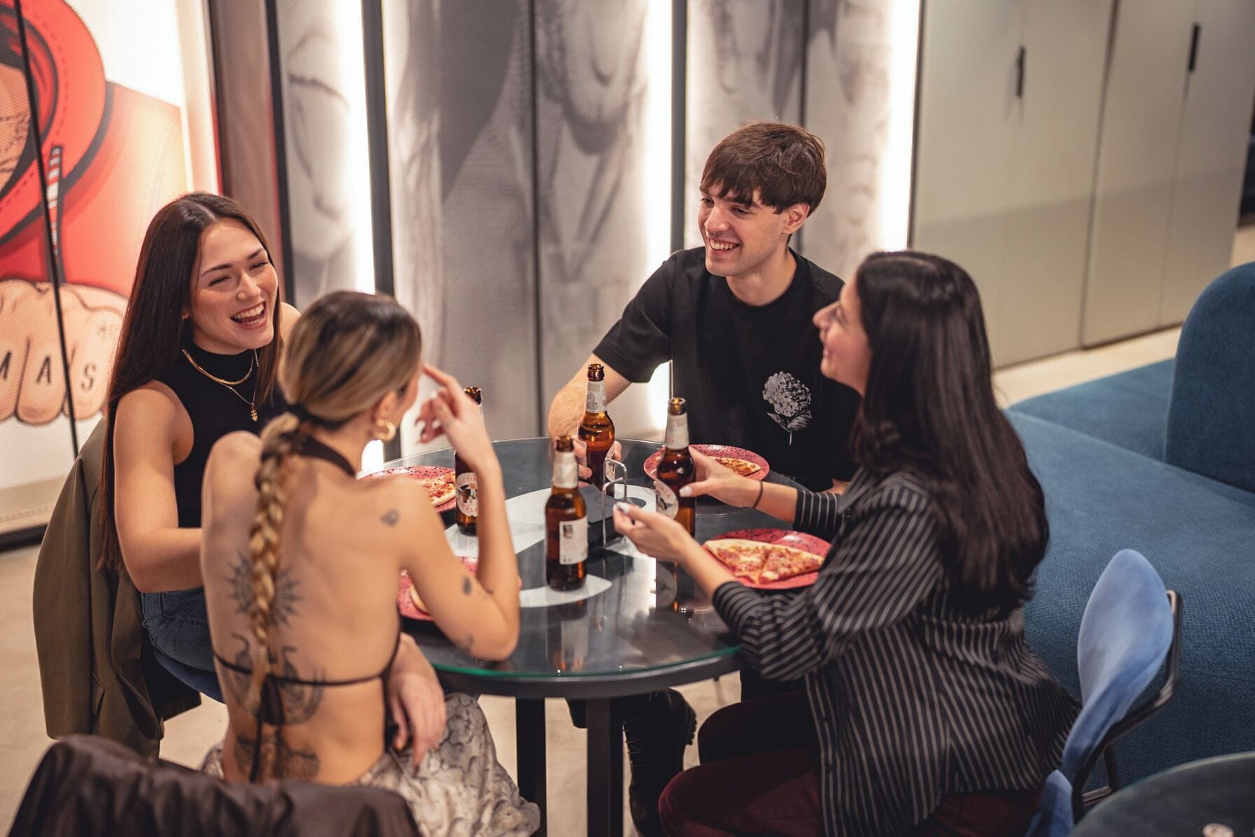 Un grupo de personas sentadas alrededor de una mesa con pizza y bebidas, en una habitación con un sofá y un cuadro grande en la pared.