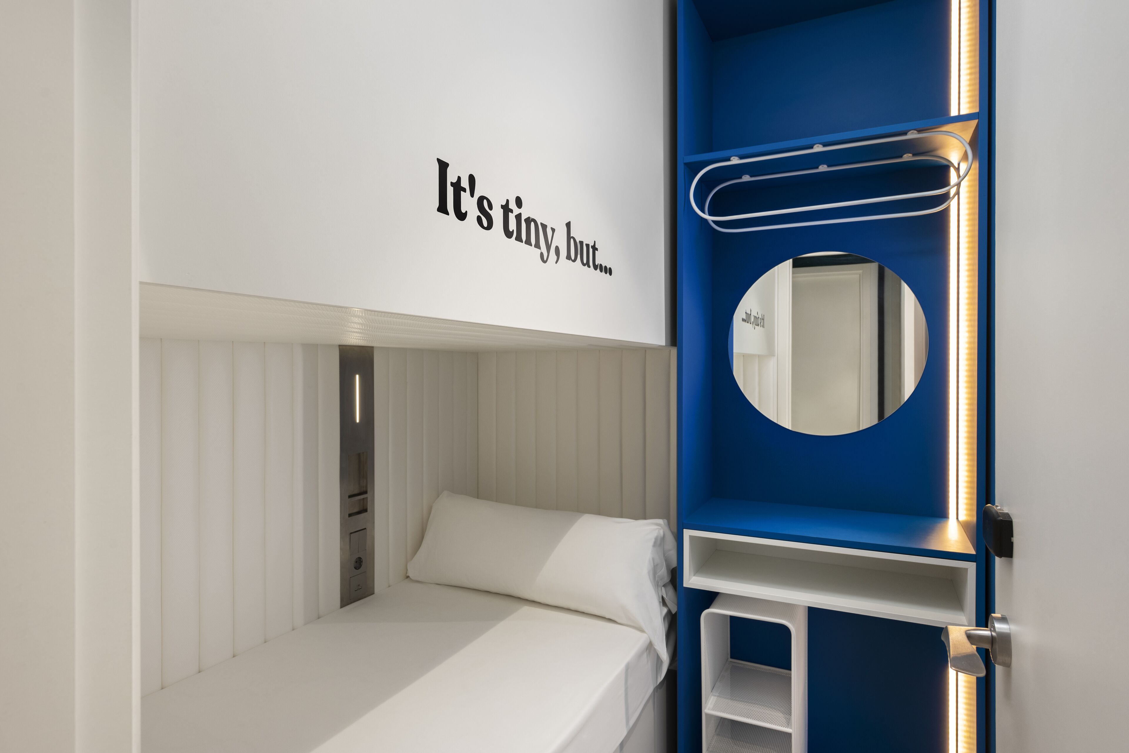 A bunk bed with a white mattress and a small shelf below. A mirror with a blue frame is mounted on the wall above the bed.