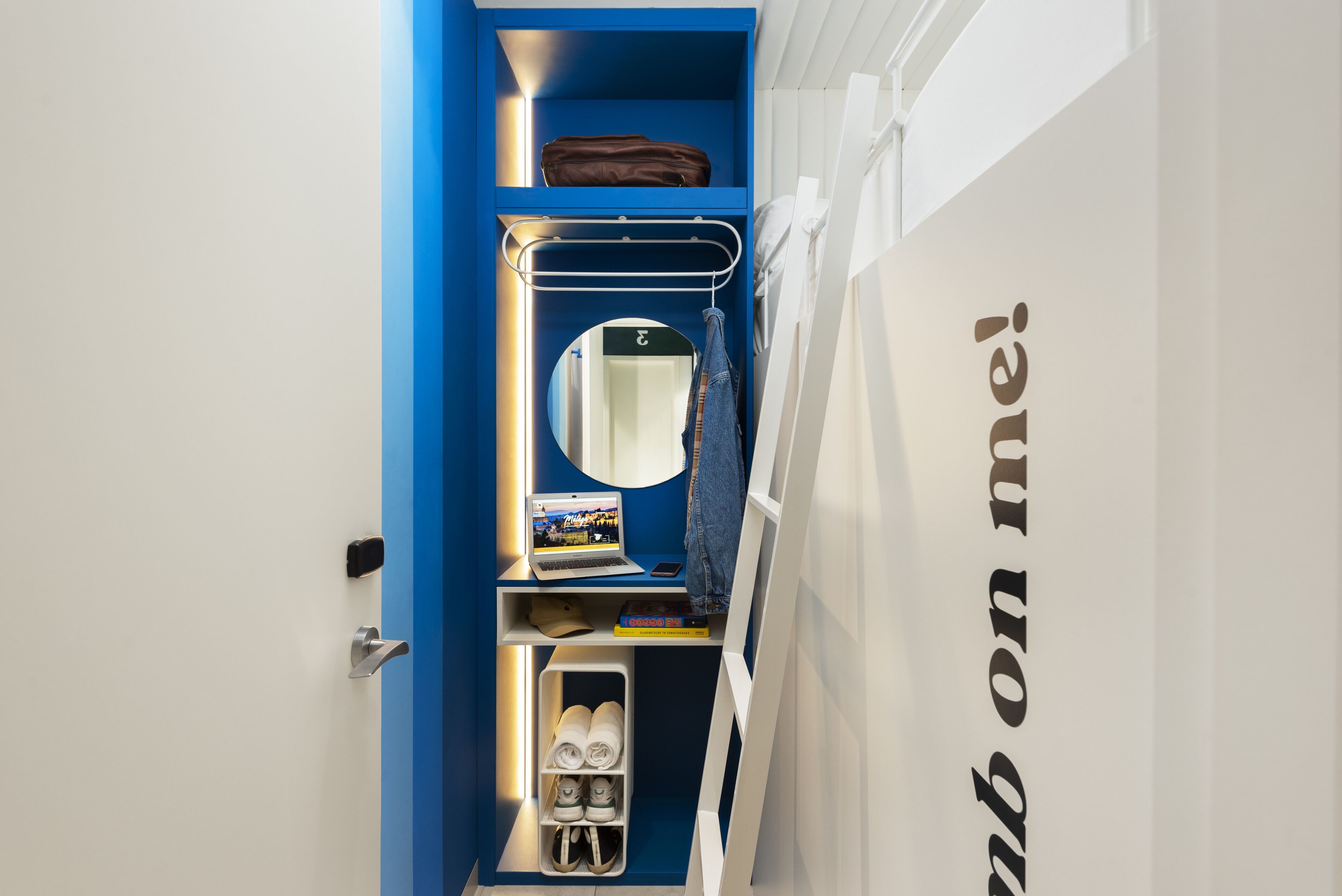 Habitación compacta con un tabique azul, una computadora portátil sobre una estantería, un espejo y un zapatero.