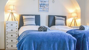 Une chambre avec deux lits, chacun recouvert d’une couverture bleue et agrémenté d’oreillers blancs. Une pancarte au-dessus des lits indique : « À bientôt à la plage ».