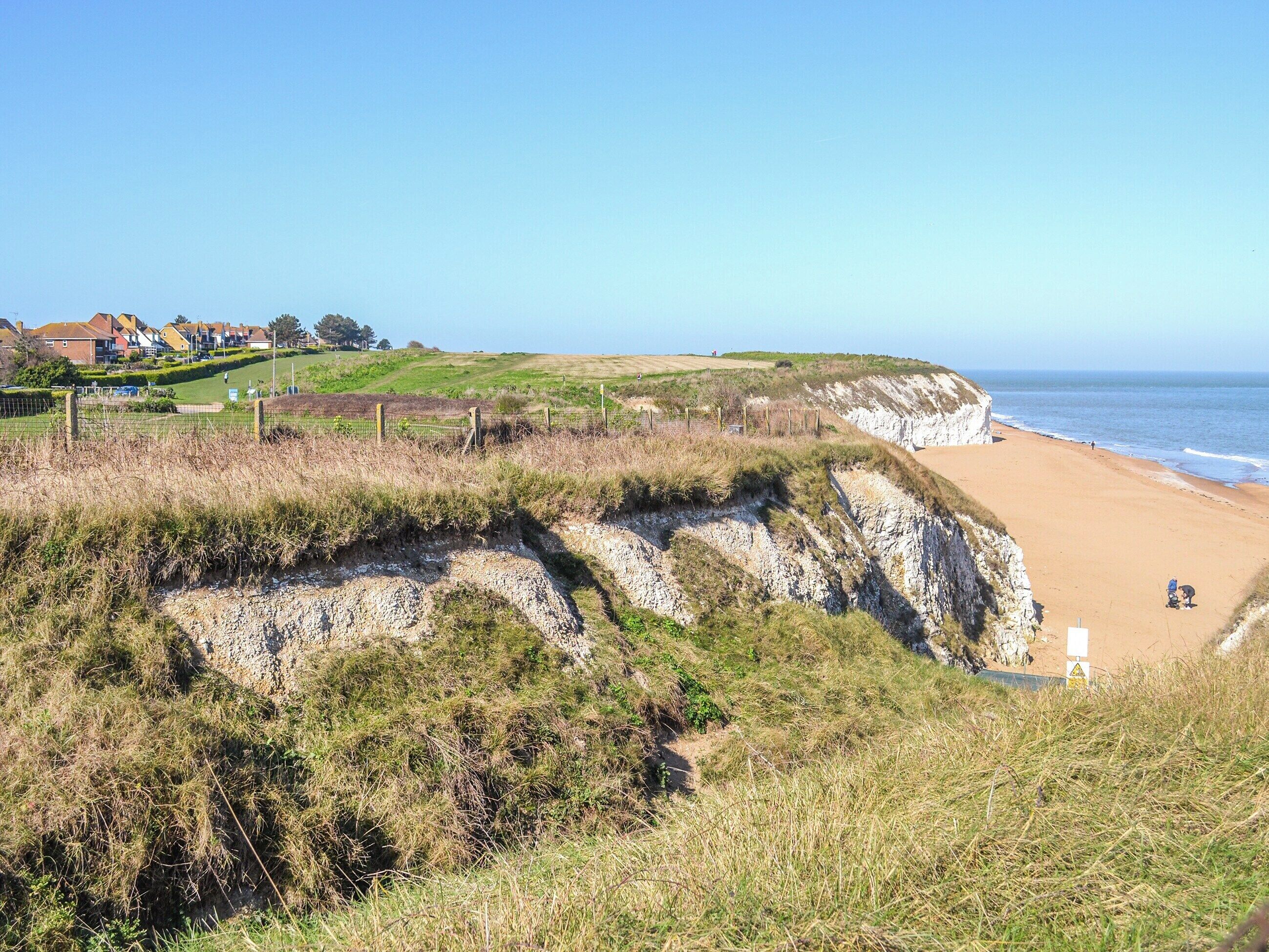 Un paisaje costero con una playa arenosa, acantilados cubiertos de hierba y algunas casas al fondo.