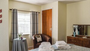 Un dormitorio con cama, una silla, una mesita con un florero y un aparador con espejo.