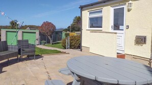 Un patio con una mesa de picnic y dos sillas de mimbre, un pequeño edificio con una ventana y una puerta verde.