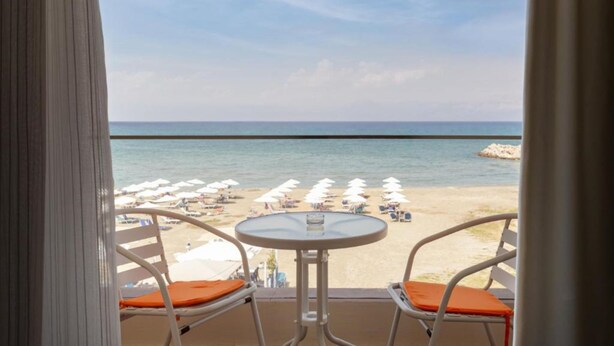 Un balcón con dos sillas blancas y una mesita pequeña, con vista a una playa con sombrillas y sillas de descanso blancas.