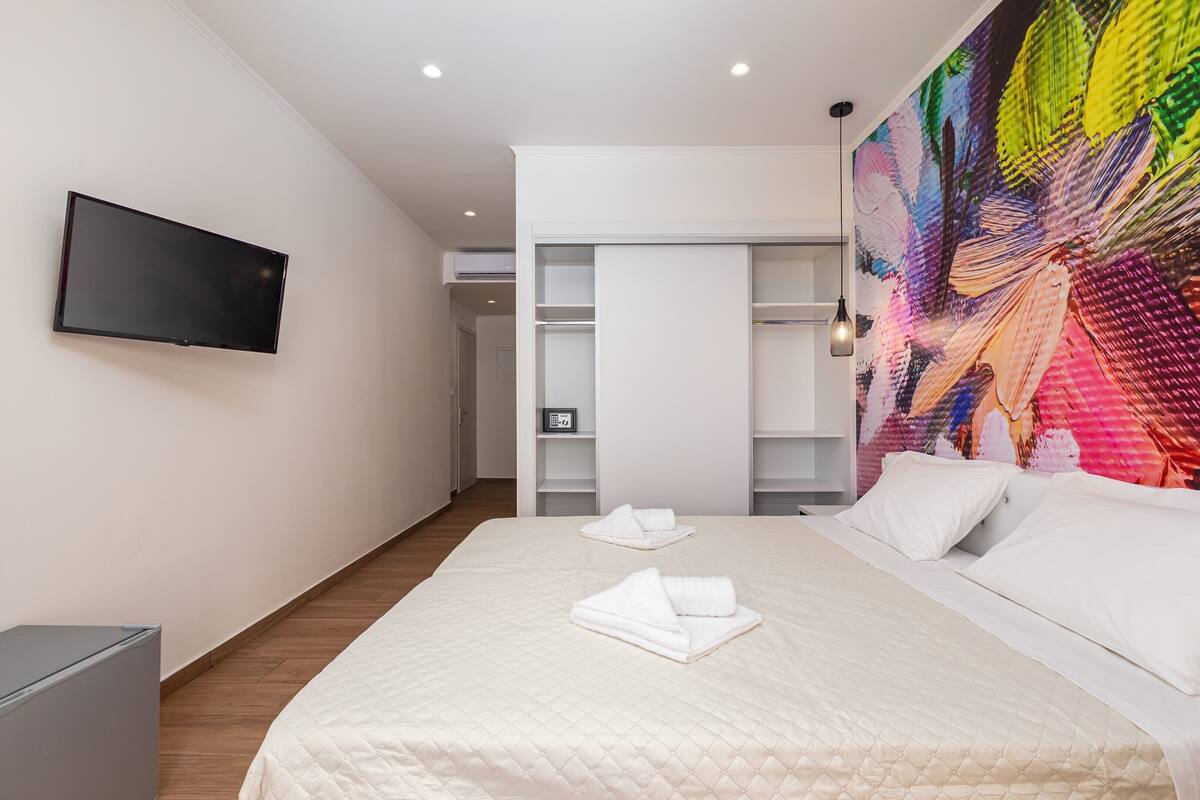 Quarto de hotel moderno com uma cama grande, televisão de tela plana, guarda-roupa e um quadro abstrato colorido na parede.