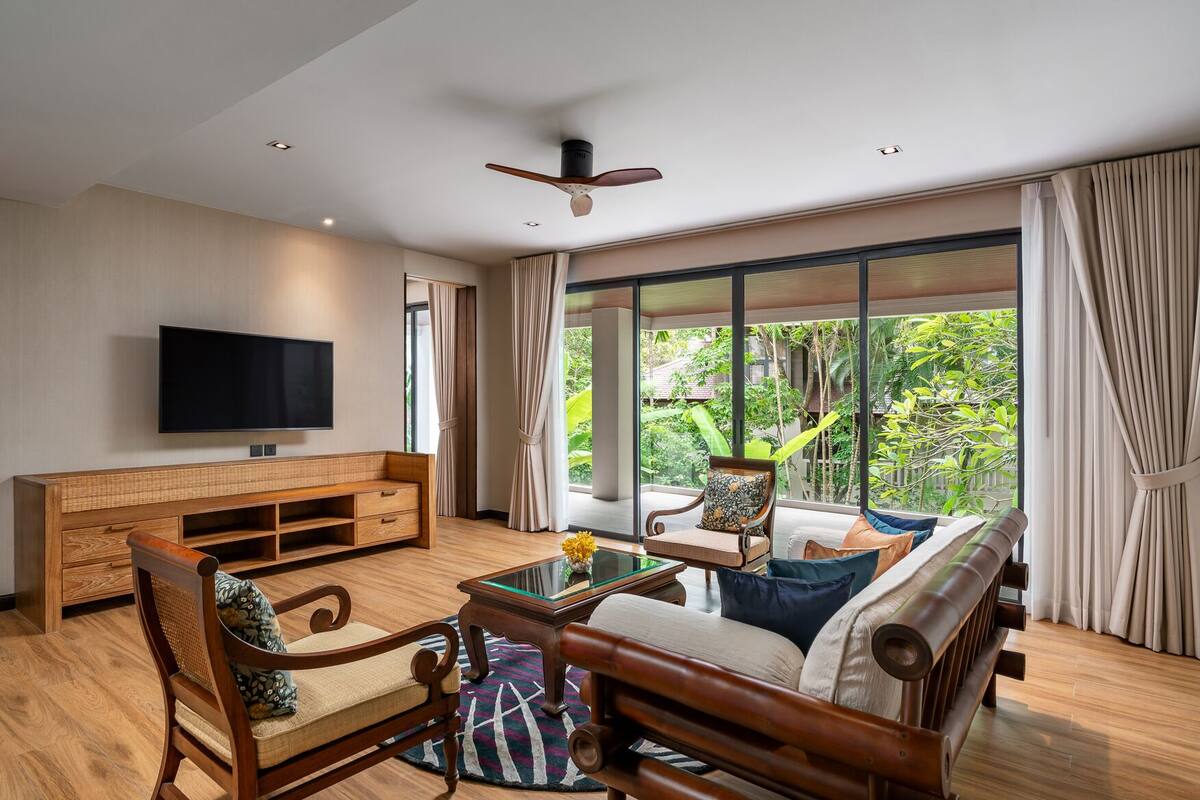 A spacious living room with a large TV, wooden furniture, a glass coffee table, and a view of lush greenery outside.