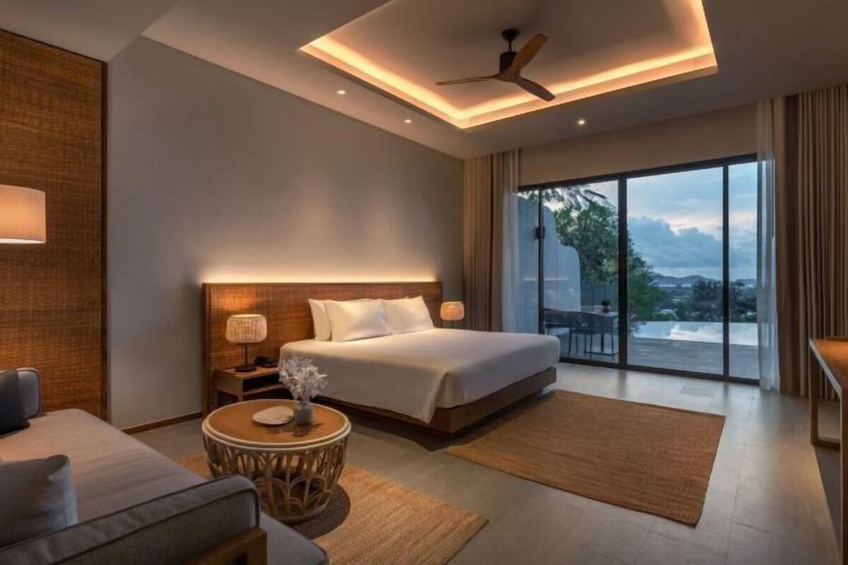 A modern hotel room with a large bed, a round side table, a ceiling fan, and a large window with a view.