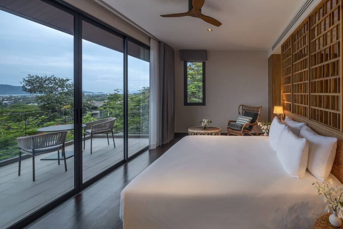A bedroom with a large bed, a view of greenery, a balcony with chairs, and a wooden wall panel.