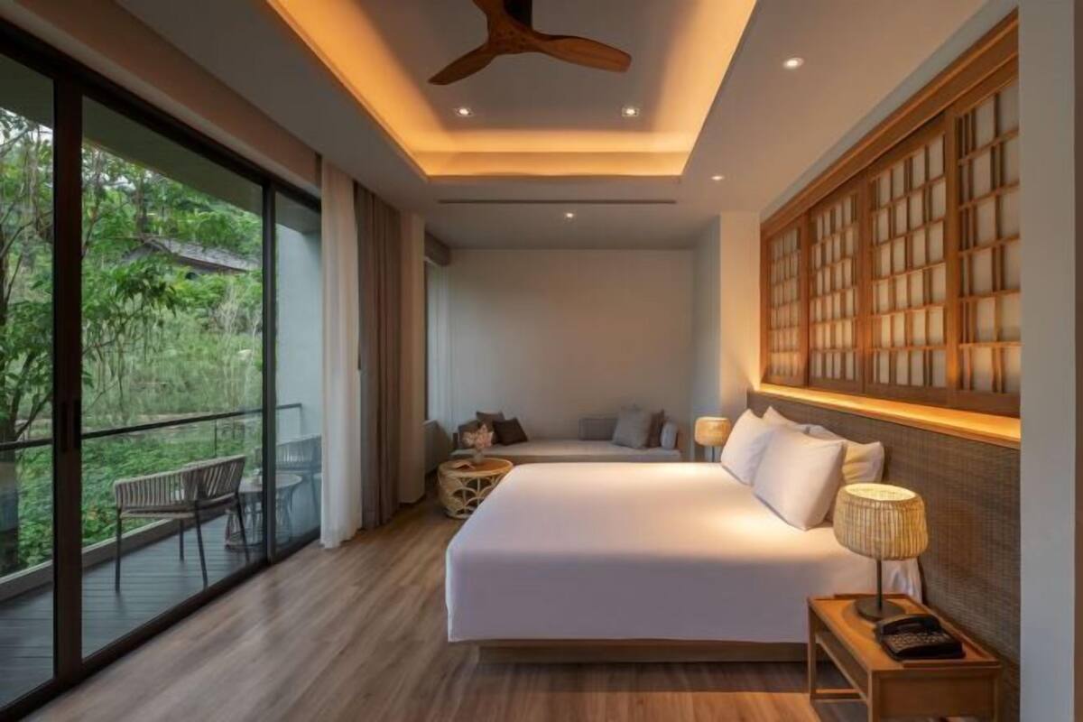 A modern hotel room with a large bed, a balcony with outdoor furniture, and a view of greenery.