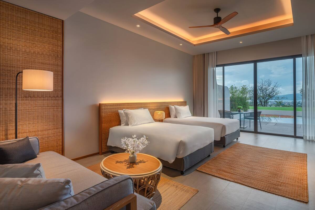 A modern hotel room with two beds, a sitting area, a dining table, and a large window offering a view of the outdoors.