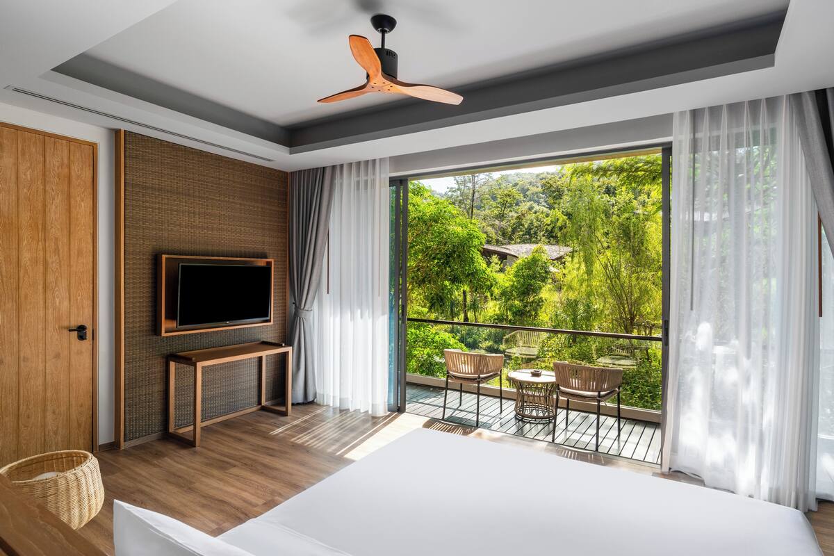 A modern hotel room with a large bed, a flat-screen TV, a wooden wardrobe, a balcony with a view of greenery, and a ceiling fan.
