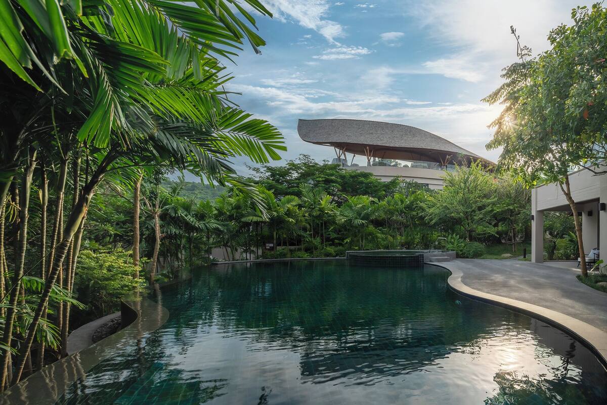 A pool surrounded by lush greenery, a modern building with a unique roof structure, and a clear sky.