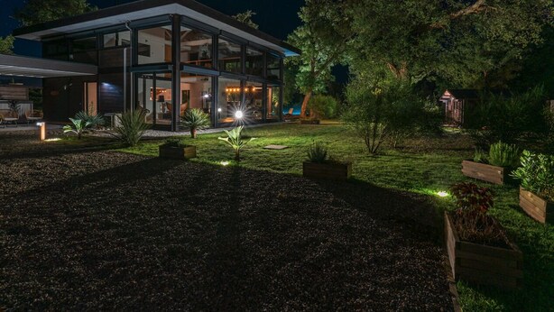 Een modern huis met een glazen gevel, een goed verlicht buitengebied en een tuin met diverse planten.