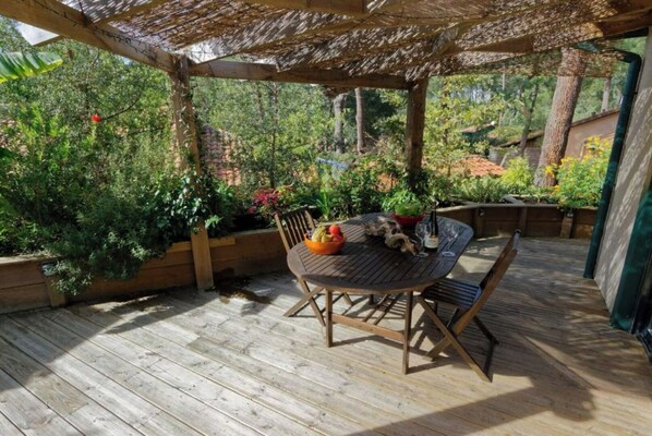 An outdoor wooden deck with a round table and chairs, surrounded by lush greenery and a pergola overhead. - Villa under the pines in Moliets (Moliets-et-Maa)