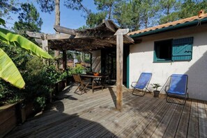 A wooden deck with a table and chairs, surrounded by lush greenery and a white building with a green window. - Villa under the pines in Moliets (Moliets-et-Maa)