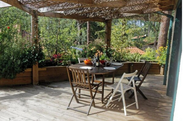 An outdoor wooden deck with a table set for dining, surrounded by lush greenery and a pergola overhead.