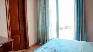 A bedroom with a wooden door, a bed with a blue bedspread, and a window with curtains. - Hotel Lindaraja (Albolote)