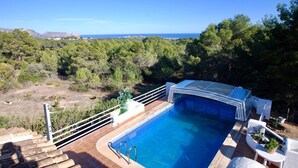 A swimming pool with a glass enclosure, surrounded by a wooden deck and white outdoor furniture. - The Great Luxury Villa Villa of Atenea (Cautivador)