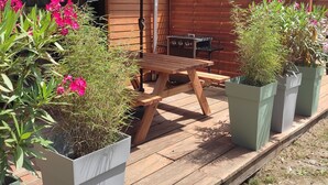 Eine Holzterrasse mit Picknicktisch und Sonnenschirm, umgeben von Kübelpflanzen und einem Grill.