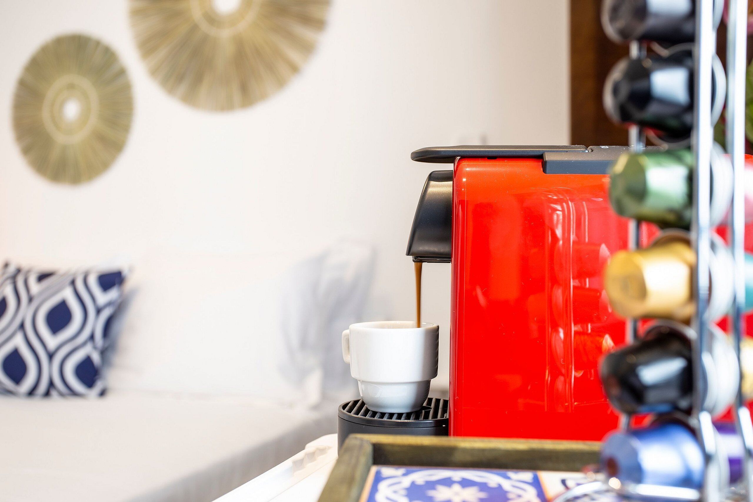 A coffee machine with a cup of coffee being dispensed, a decorative wall plate, and a patterned pillow on a bed.