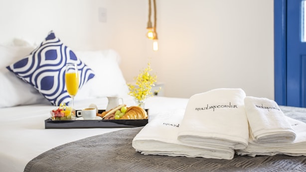 A neatly made bed with a tray of breakfast, including croissants, fruit, and a glass of orange juice. There are folded towels with "Brazilian Corner" branding and a small vase with yellow flowers. - Vila Grega (São Paulo)