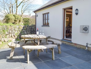 A round wooden table with benches, a bottle of wine, and two wine glasses on a patio with stone walls and a stone pathway.