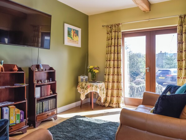 A living room with a bookshelf, a table with a floral tablecloth, a sofa, and a large window with curtains.
