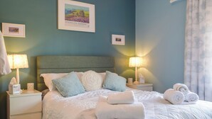 A neatly made bed with pillows and rolled towels, a bedside table with a lamp, and a framed picture on the wall.
