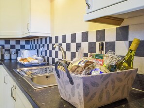 Une cuisine avec un dosseret à carreaux, un évier et un panier contenant du pain, une bouteille et d’autres produits alimentaires.