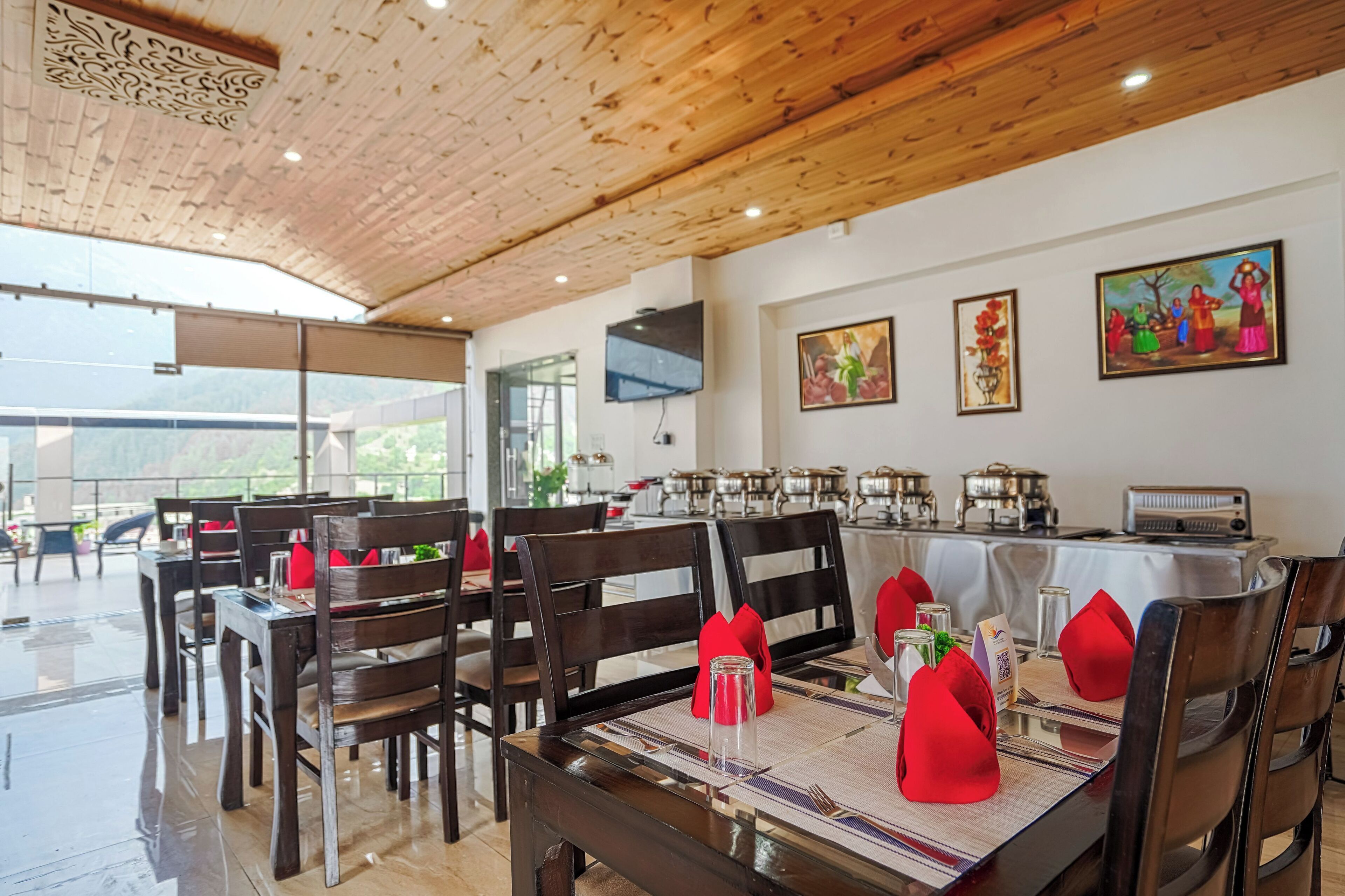 A restaurant with wooden ceiling and walls, tables set with red napkins, and paintings on the wall.