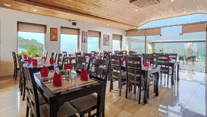 Un comedor amplio con techo de madera y ventanas grandes que ofrecen vistas del exterior. Las mesas están cuidadosamente preparadas con servilletas rojas y cristalería.
